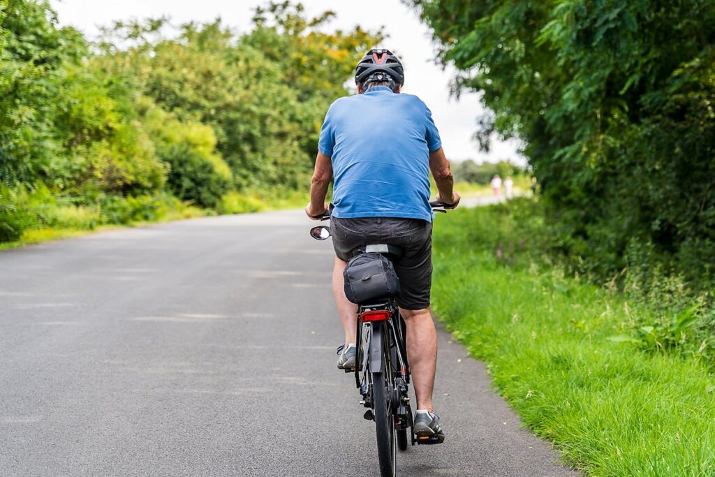 Uomo maturo in bicicletta su strada panoramica, simbolo di resistenza fisica e benessere cardiovascolare.