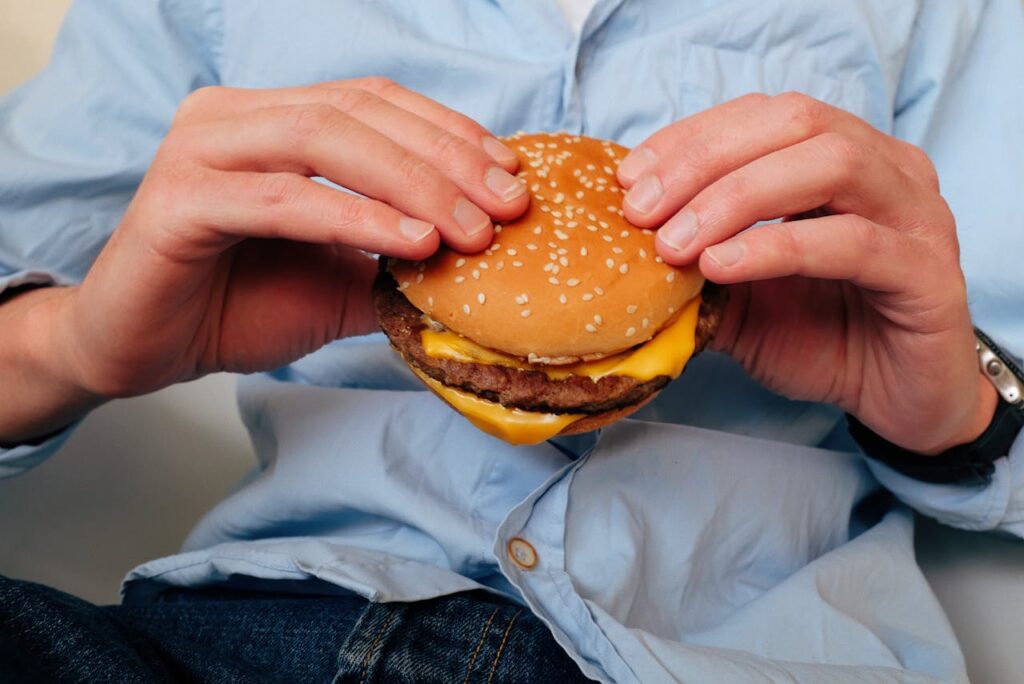 Uomo adulto che tiene un hamburger fast food, esempio di alimenti che fanno male alla salute se consumati frequentemente.